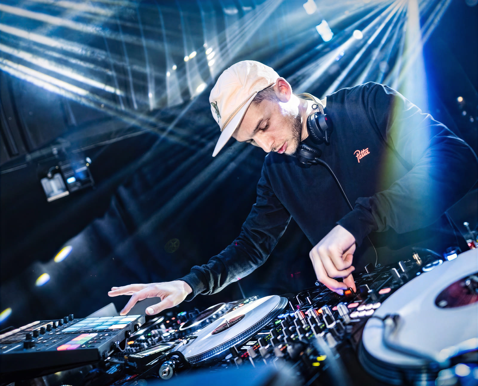 Rafik performing on two turntables and a mixer, one hand on the platter and one on the controls, stage beams behind him.
